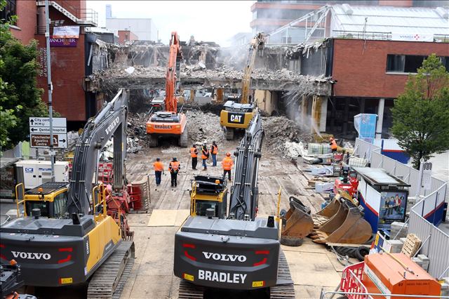 Demolition site with excavators on timber mats for protecting the ground tearing down a large brick building, surrounded by workers and equipment.